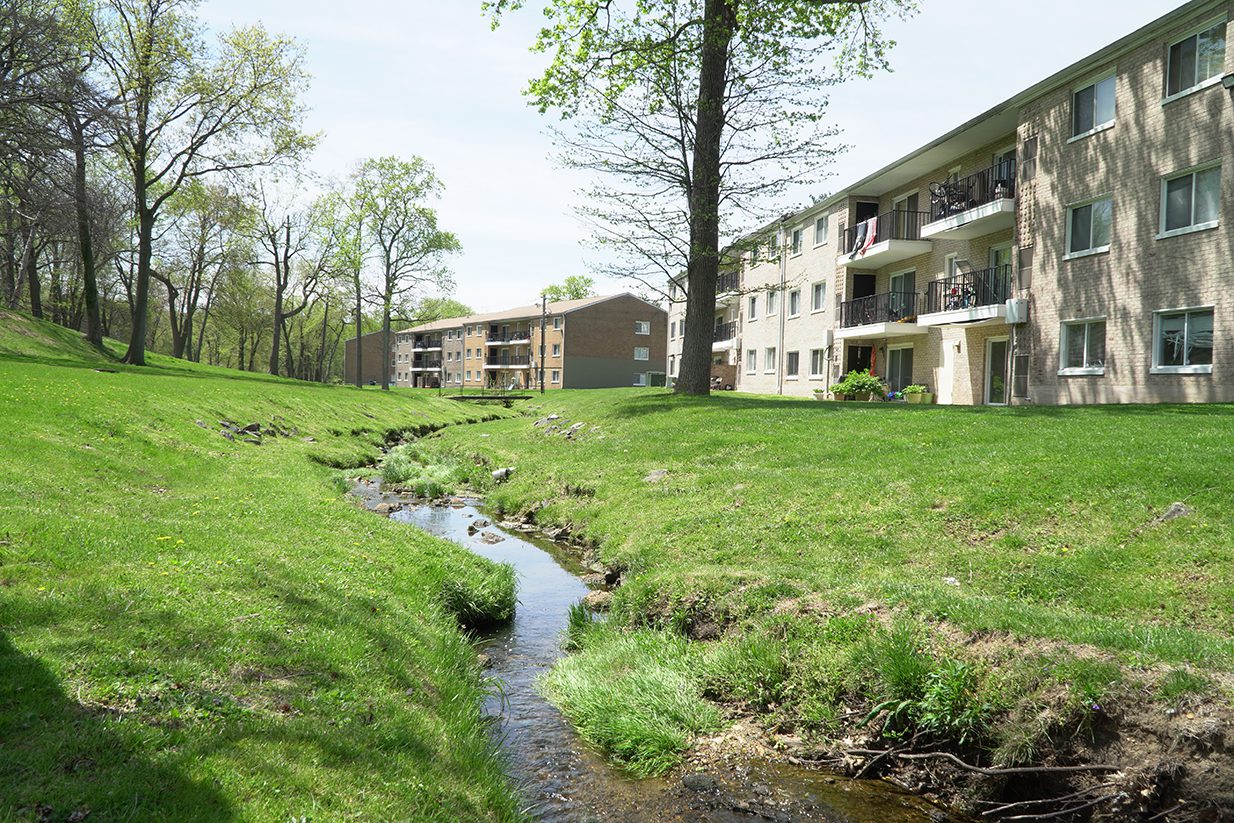 A serene North Hills apartment complex with brick buildings overlooking a small, winding creek, surrounded by lush green grass and scattered trees on a sunny day.